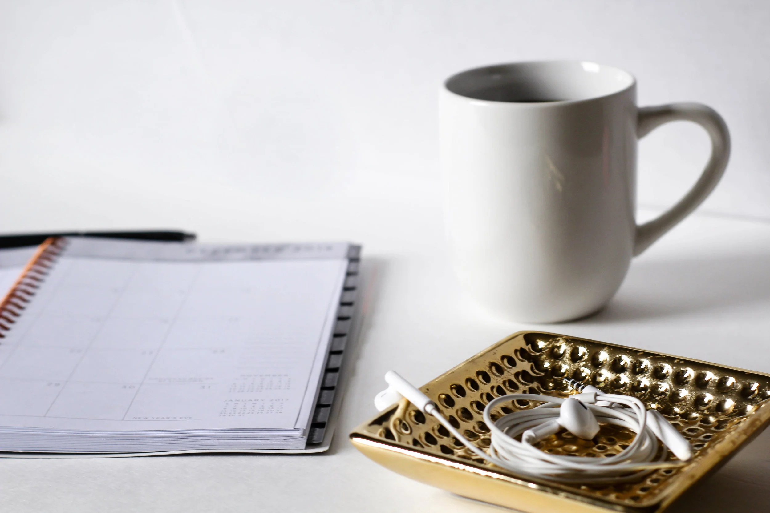 Flat lay of a white mug, a spiral planner with a pen, and white earbuds on a gold decorative tray, clean minimal mood
