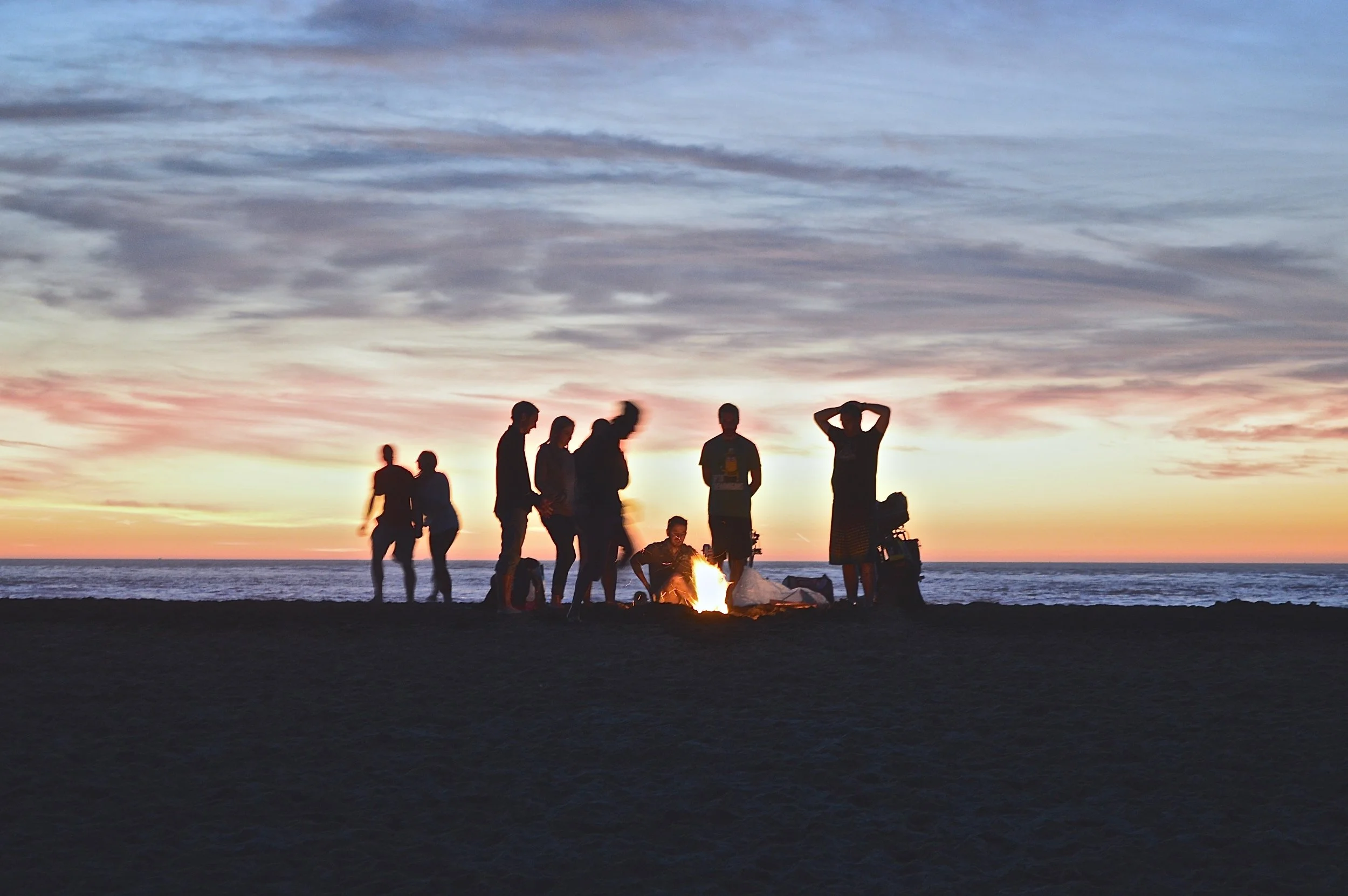 Silhouettes of people gathered around a glowing bonfire at sunset, warm orange and blue tones, relaxed social mood