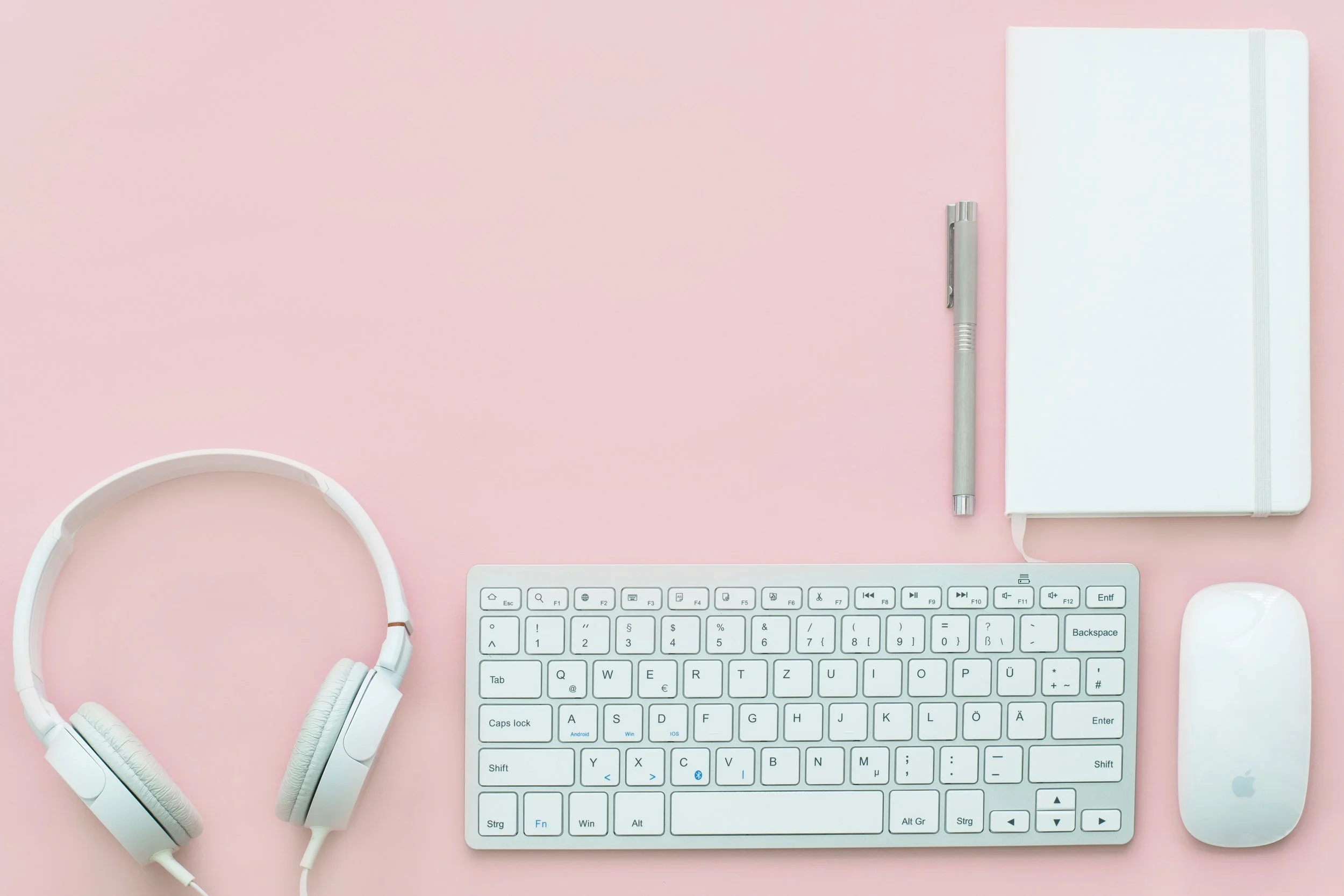 Flat lay of white headphones, keyboard, mouse, pen, and notebook on a pink background, minimalist clean mood