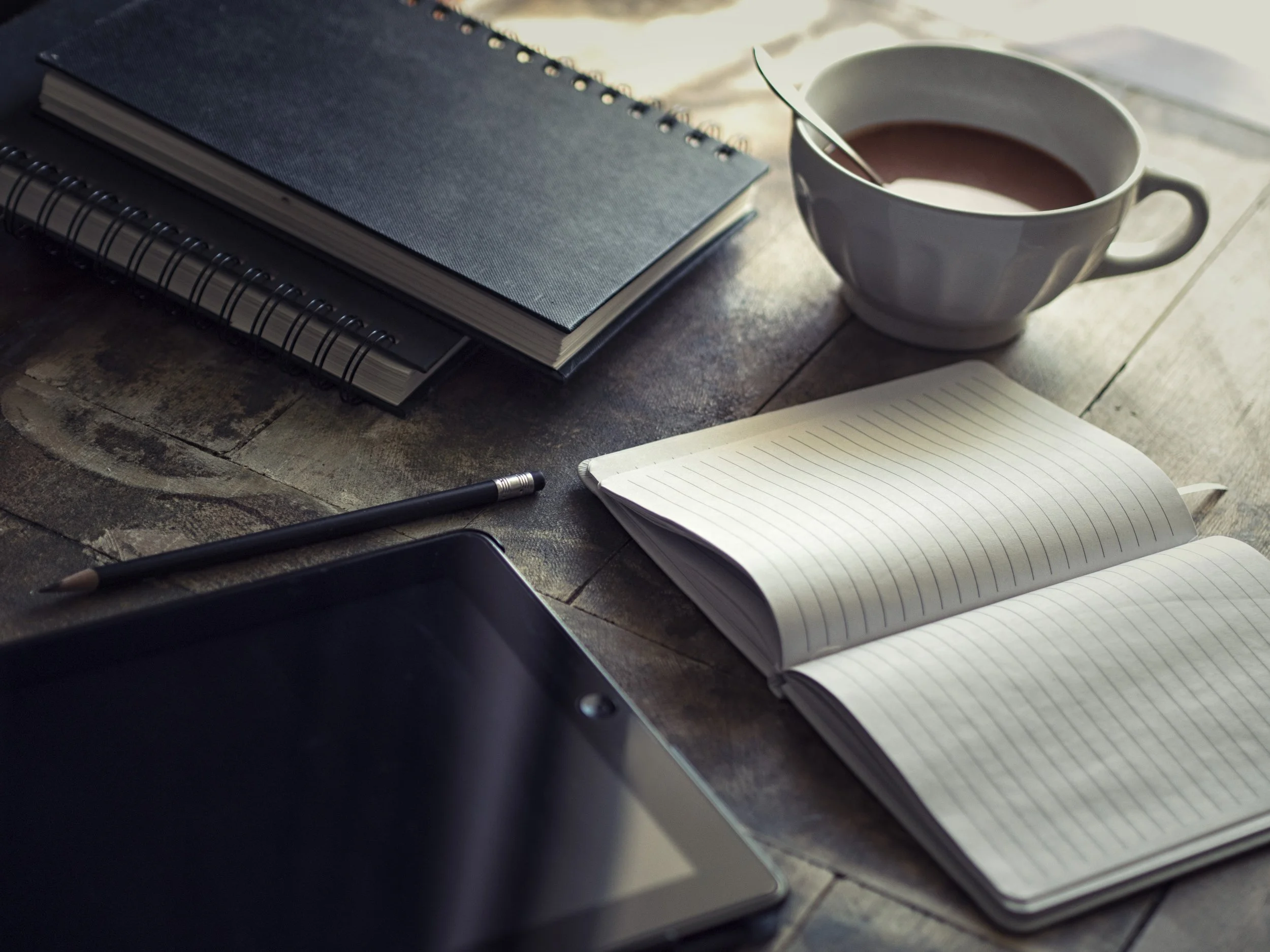 Notebooks, a pencil, a tablet, and a cup of tea arranged on a wooden table, calm neutral tones